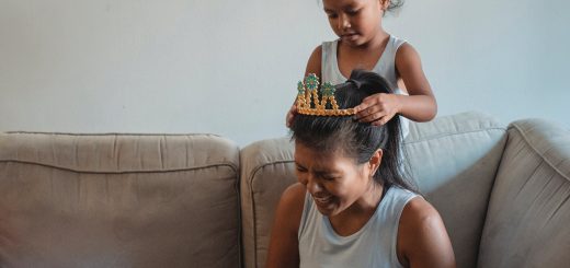 Adorable Asian girl putting crown on laughing mothers head siting on cozy couch and working on laptop at home